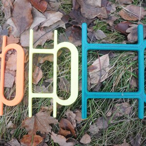Könnte beinhalten: Drei bunte Plastikbuchstaben, die das Wort "HOP" in Orange, Gelb und Türkis buchstabieren. Die Buchstaben sind in einer Reihe auf einem Bett aus braunen Blättern und grünem Gras angeordnet.