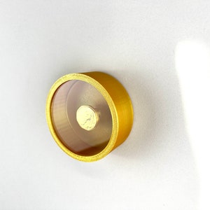 Könnte beinhalten: Eine runde, goldfarbene Box mit einem durchsichtigen Deckel. Der Deckel ist geöffnet und zeigt ein kleines, goldfarbenes Objekt im Inneren.