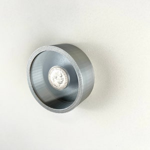 Könnte beinhalten: Eine Silbermünze wird in einem runden, silberfarbenen Rahmen mit einer durchsichtigen Plastikabdeckung ausgestellt. Der Rahmen ist an einer weißen Wand befestigt.