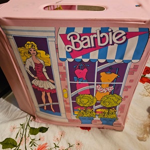 May include: Pink plastic Barbie playset shaped like a storefront with a blue and white awning that says "Barbie". The storefront has a window display with two dresses and two potted plants. A Barbie doll is standing in the doorway.