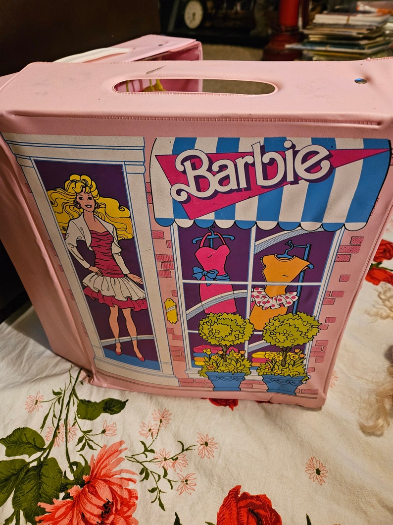 May include: Pink plastic Barbie playset shaped like a storefront with a blue and white awning that says "Barbie". The storefront has a window display with two dresses and two potted plants. A Barbie doll is standing in the doorway.