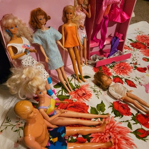 May include: A group of dolls, including Barbie dolls, are arranged on a floral tablecloth. The dolls are wearing various outfits, including a striped dress, a yellow dress, and a swimsuit. There is a pink suitcase with a built-in closet in the background.