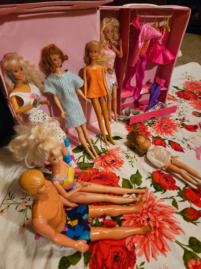 May include: A group of dolls, including Barbie dolls, are arranged on a floral tablecloth. The dolls are wearing various outfits, including a striped dress, a yellow dress, and a swimsuit. There is a pink suitcase with a built-in closet in the background.