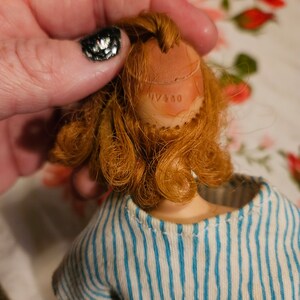 May include: A vintage doll with red hair and a blue and white striped shirt. The doll's head has a brown plastic cap with the text "NY 450" printed on it.
