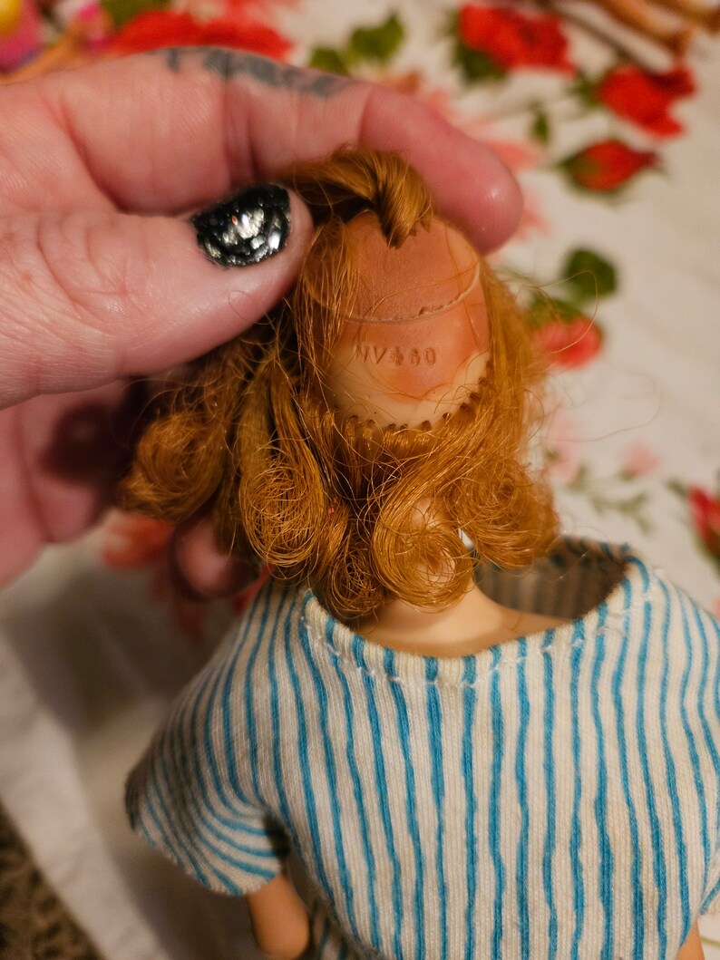 May include: A vintage doll with red hair and a blue and white striped shirt. The doll's head has a brown plastic cap with the text "NY 450" printed on it.