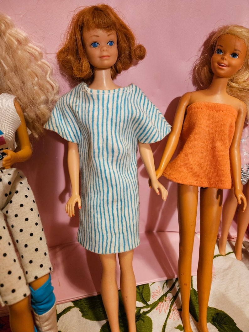 May include: Three vintage Barbie dolls with different hairstyles and outfits. The doll on the left is wearing a white shirt with black polka dots and blue pants. The doll in the center is wearing a blue and white striped dress. The doll on the right is wearing a peach-colored top and a white skirt.