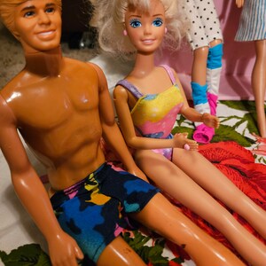 May include: A vintage Barbie and Ken doll couple wearing swimwear. The Barbie doll is wearing a yellow and pink swimsuit with a floral pattern. The Ken doll is wearing blue, green, and pink swim trunks. The dolls are sitting on a floral patterned fabric.