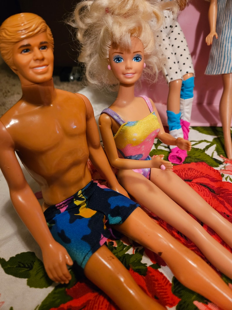 May include: A vintage Barbie and Ken doll couple wearing swimwear. The Barbie doll is wearing a yellow and pink swimsuit with a floral pattern. The Ken doll is wearing blue, green, and pink swim trunks. The dolls are sitting on a floral patterned fabric.