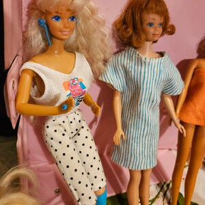 May include: Three vintage Barbie dolls with different hairstyles and outfits. The doll on the left has blonde hair, a white tank top with a colorful graphic, and black and white polka dot pants. The doll in the center has brown hair, a blue and white striped dress, and blue eyes. The doll on the right has blonde hair, a pink dress, and blue eyes.