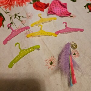 May include: Five colorful plastic hangers with a decorative design and a purple and rainbow feather boa on a white floral tablecloth.