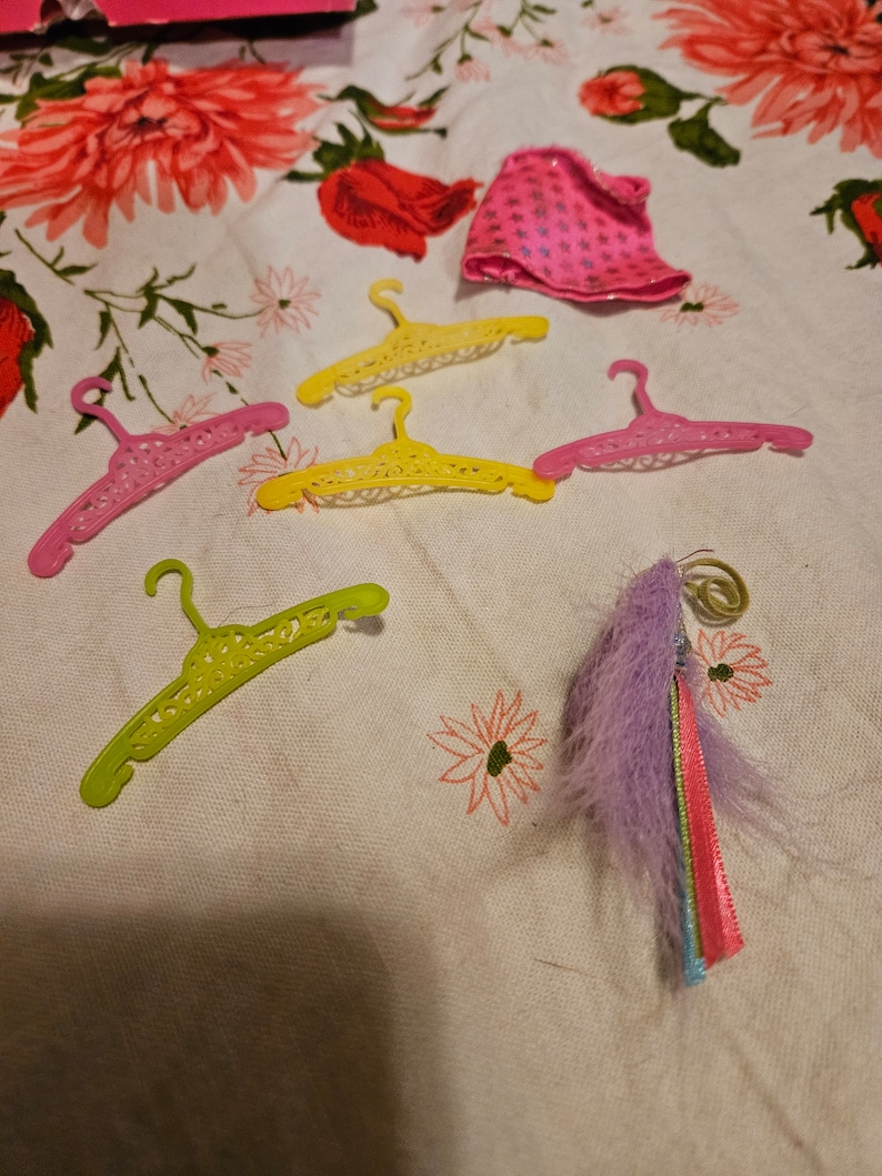 May include: Five colorful plastic hangers with a decorative design and a purple and rainbow feather boa on a white floral tablecloth.