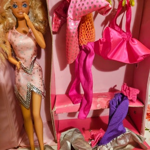 May include: A pink plastic dollhouse with a blonde doll wearing a pink dress with silver stars. The dollhouse has a pink interior with a closet that has several pink and purple outfits hanging on hangers.