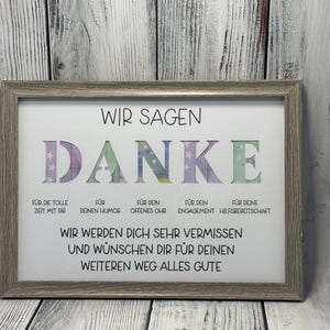 May include: A framed print with the word "DANKE" in colorful letters, surrounded by stars. The text below reads "Wir sagen Danke" and includes reasons for thanking someone, such as "for your humor" and "for your helpfulness". The print also says "Wir werden dich sehr vermissen und w&uuml;nschen dir f&uuml;r deinen weiteren Weg alles Gute".