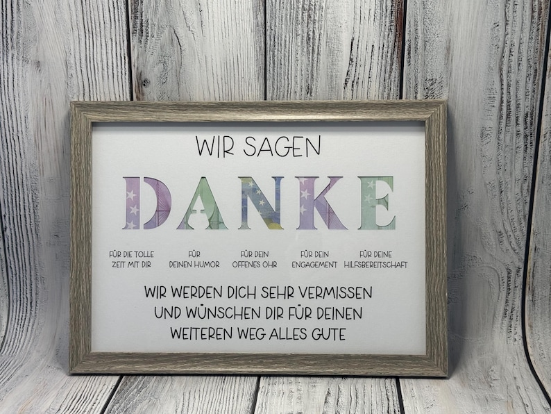 May include: A framed print with the word "DANKE" in colorful letters, surrounded by stars. The text below reads "Wir sagen Danke" and includes reasons for thanking someone, such as "for your humor" and "for your helpfulness". The print also says "Wir werden dich sehr vermissen und w&uuml;nschen dir f&uuml;r deinen weiteren Weg alles Gute".