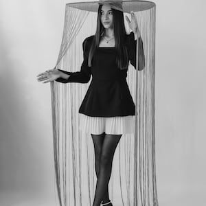 Puede incluir: Una mujer con un vestido negro con una falda plisada blanca, medias negras y tacones altos. Lleva un sombrero grande con ala ancha y una franja de hilos colgantes. El fondo es blanco liso.