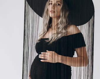 May include: A pregnant woman wearing a black off-the-shoulder dress and a large black hat with fringe. The hat is covering her face and the fringe is hanging down around her.