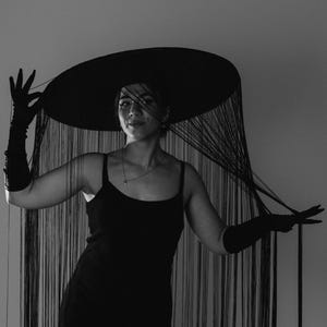 Puede incluir: Una persona con un vestido negro y un sombrero de ala ancha con largas franjas negras. El sombrero cubre la cara de la persona.