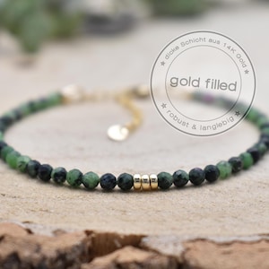 Könnte beinhalten: Armband mit grünen und schwarzen Edelsteinen und goldgefüllten Akzenten. Das Armband hat ein zartes Design mit kleinen, runden Perlen und einer goldenen Kette. Der Text "gold filled" ist in einer kreisförmigen Grafik sichtbar.