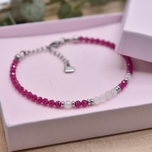 Könnte beinhalten: Ein rosa-weißes Perlenarmband mit einem silbernen Herz-Anhänger. Das Armband befindet sich in einer rosa Box.
