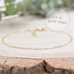 Könnte beinhalten: Ein zartes, goldgefülltes Kettenarmband mit einem schlichten, eleganten Design. Das Armband wird auf einer rustikalen Holzoberfläche präsentiert, mit einem verschwommenen Hintergrund aus Grün. Der Text "14K gold filled" befindet sich in einem kreisförmigen Rahmen.