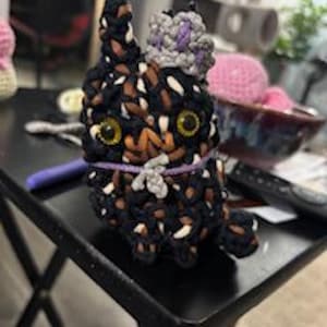 May include: A handmade crochet cat doll with yellow eyes, black, brown, and white yarn, and a small purple and gray hat. The cat is sitting on a black table. Other crochet items are visible in the background.