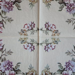 Op de afbeelding: Een witte papieren servet met een bloemenpatroon. Het ontwerp bevat roze, paarse en witte bloemen met groene bladeren. De bloemen zijn symmetrisch gerangschikt.