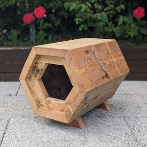 May include: A hexagonal wooden pet bed with a large opening for entry. The bed is made of reclaimed wood and has a natural finish. It is sitting on a grey concrete patio.