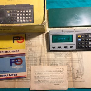 May include: A vintage Elektronika MK-52 calculator with a green display showing numbers. The image includes the calculator's box, a green case, instruction manuals, and a schematic diagram. The calculator has a grey body and a black keyboard.