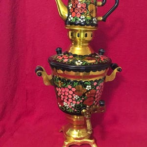 Può includere: Un samovar in metallo dorato con base e coperchio dipinti di nero, decorato con motivi floreali rossi e rosa dipinti a mano. L'unità include una teiera abbinata sulla parte superiore, due manici laterali, un rubinetto centrale e una base solida.
