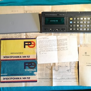 Puede incluir: Una calculadora programable Elektronika MK-52 de color gris oscuro con pantalla digital, una cubierta protectora gris, dos manuales de instrucciones con portadas amarilla y azul, una hoja informativa blanca y un gran esquema técnico.