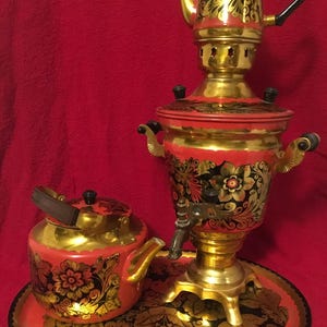 May include: A traditional Russian samovar set with a teapot and serving tray. The set features a red and gold floral design with black accents. The samovar has a spigot and sits on a decorative base. The teapot has a wooden handle.