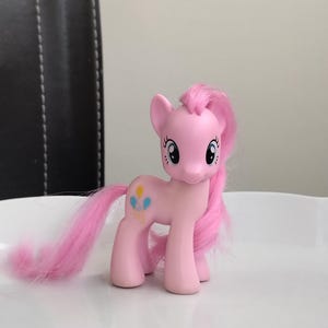 May include: A pink plastic pony figurine with a long, flowing pink mane and tail. The pony has large, expressive blue eyes and a cutie mark on its flank featuring three colorful balloons in yellow, blue, and pink. The figure stands on four legs.