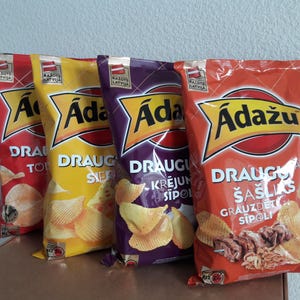 Op de afbeelding: Vier zakken aardappelchips van het merk Adažu in rode, gele, paarse en oranje verpakkingen. Elke zak heeft de merknaam in een gele banner. De zakken tonen verschillende smaken, met tekst in het Lets.