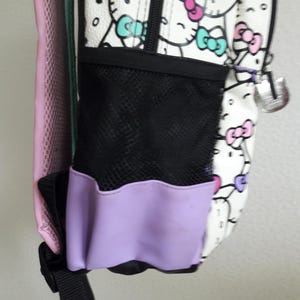 Puede incluir: Mochila blanca con un estampado repetido de personajes de dibujos animados con lazos rosas, morados y turquesas. Tiene un bolsillo lateral de malla negra con ribete lavanda y un charm plateado. La parte trasera de la mochila es rosa.