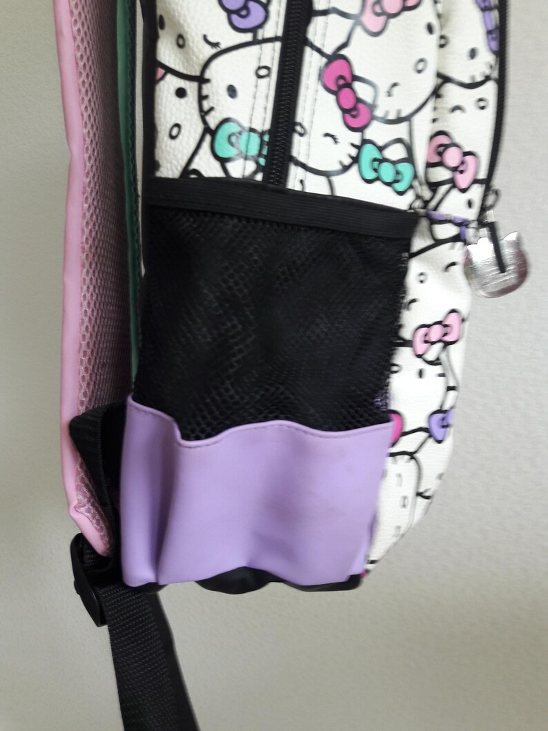 Puede incluir: Mochila blanca con un estampado repetido de personajes de dibujos animados con lazos rosas, morados y turquesas. Tiene un bolsillo lateral de malla negra con ribete lavanda y un charm plateado. La parte trasera de la mochila es rosa.
