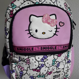 Puede incluir: Una mochila de Hello Kitty rosa y blanca con ribete negro. La parte delantera presenta una gran cara de Hello Kitty con un lazo rosa brillante. La mochila tiene un patr&oacute;n repetido de caras y lazos de Hello Kitty. La palabra "Smiggle" est&aacute; impresa en una banda rosa.