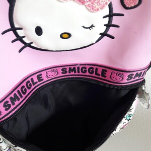 Puede incluir: Mochila rosa de Hello Kitty con lazo brillante y detalle de coraz&oacute;n. La mochila tiene un interior negro y cierre de cremallera. La palabra "Smiggle" est&aacute; impresa en una banda rosa en la parte superior.