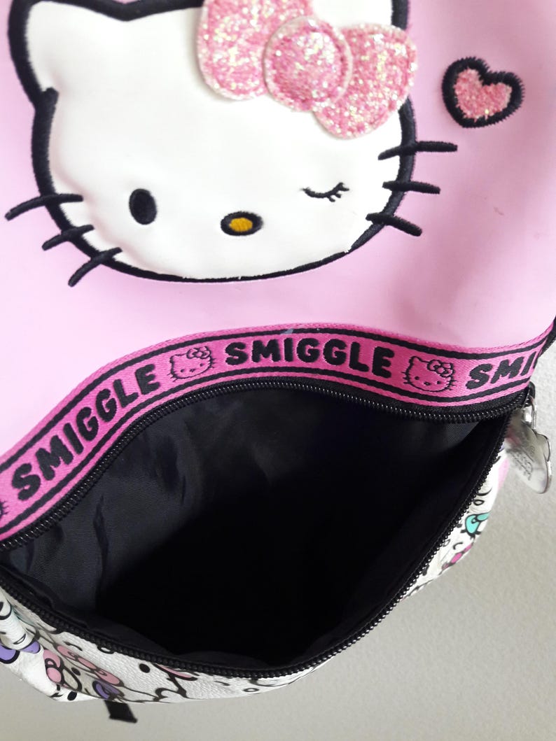 Puede incluir: Mochila rosa de Hello Kitty con lazo brillante y detalle de coraz&oacute;n. La mochila tiene un interior negro y cierre de cremallera. La palabra "Smiggle" est&aacute; impresa en una banda rosa en la parte superior.