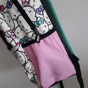 Puede incluir: Una mochila blanca con un patr&oacute;n repetido de caras y lazos de Hello Kitty en rosa, morado y turquesa. La mochila tiene un bolsillo lateral rosa, una cremallera negra y una correa negra. El tirador de la cremallera dice "Hello Kitty".
