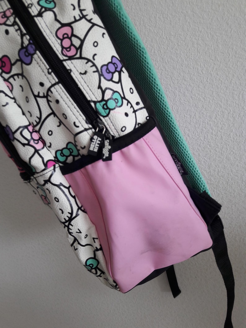 Puede incluir: Una mochila blanca con un patr&oacute;n repetido de caras y lazos de Hello Kitty en rosa, morado y turquesa. La mochila tiene un bolsillo lateral rosa, una cremallera negra y una correa negra. El tirador de la cremallera dice "Hello Kitty".