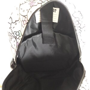 Puede incluir: Mochila blanca con interior negro, con un patr&oacute;n repetido de personajes de Hello Kitty y dise&ntilde;os florales en rosa, morado y negro. El texto "HELLO KITTY" es visible en una etiqueta blanca en el interior.