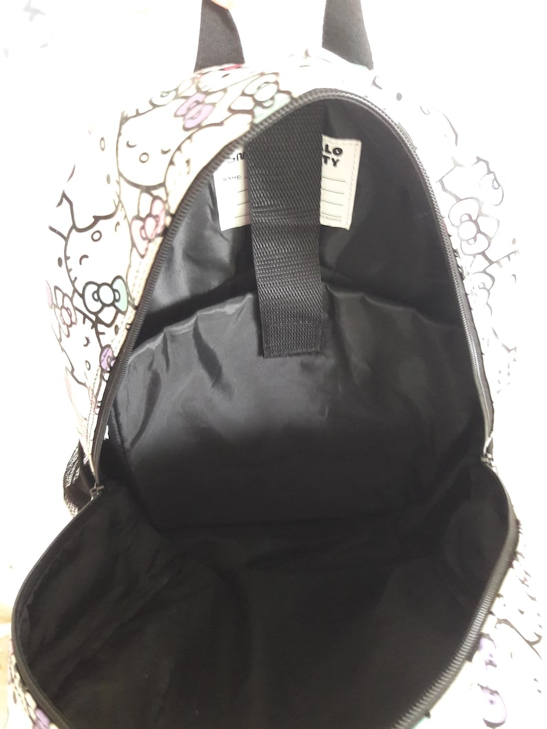 Puede incluir: Mochila blanca con interior negro, con un patr&oacute;n repetido de personajes de Hello Kitty y dise&ntilde;os florales en rosa, morado y negro. El texto "HELLO KITTY" es visible en una etiqueta blanca en el interior.