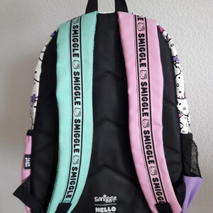 Puede incluir: Mochila negra con correas verde menta y rosa. Las correas tienen la palabra "Smiggle" impresa. La mochila presenta un dise&ntilde;o de Hello Kitty y el logotipo "Smiggle Hello Kitty". La mochila tiene una parte trasera negra y un bolsillo lateral morado.