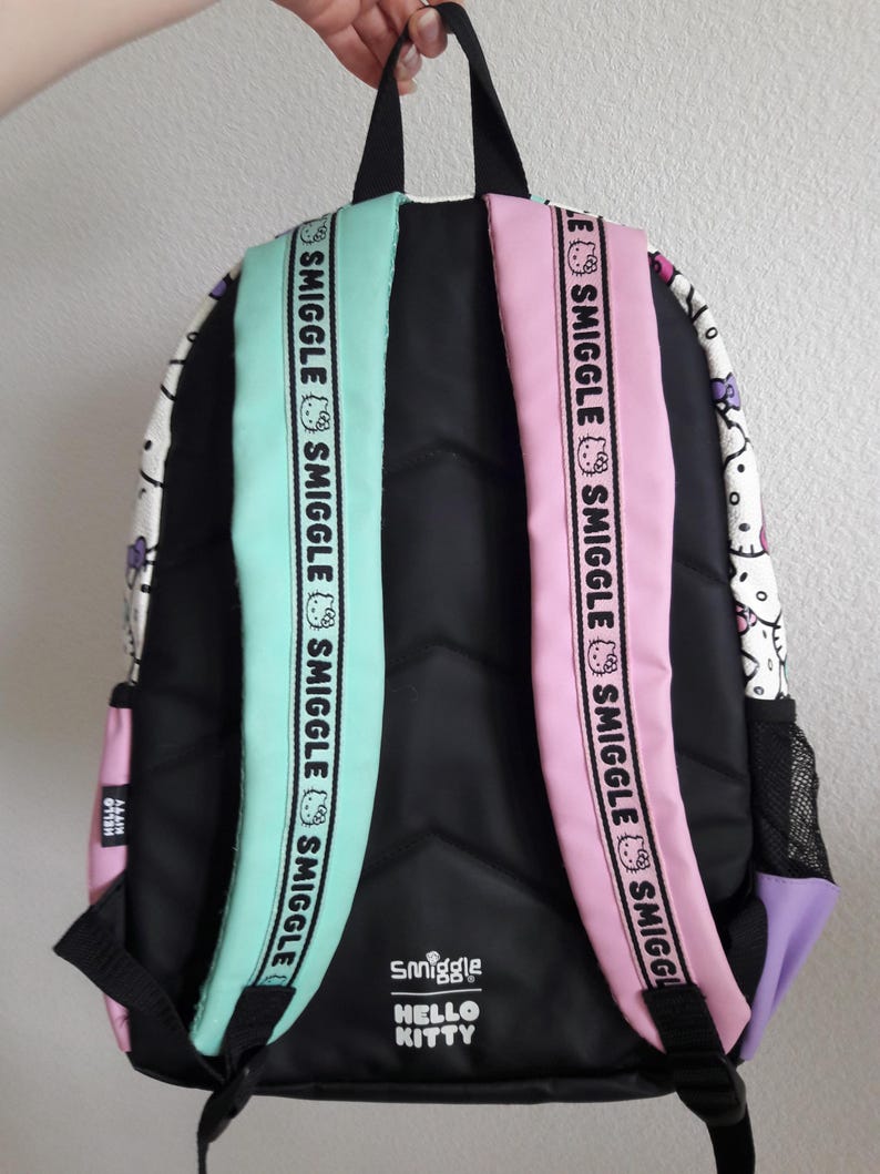Puede incluir: Mochila negra con correas verde menta y rosa. Las correas tienen la palabra "Smiggle" impresa. La mochila presenta un dise&ntilde;o de Hello Kitty y el logotipo "Smiggle Hello Kitty". La mochila tiene una parte trasera negra y un bolsillo lateral morado.