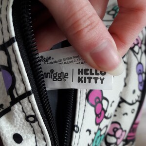 Puede incluir: Bolso blanco con cremallera negra y dise&ntilde;o de Hello Kitty. El bolso presenta un patr&oacute;n repetido de caras y lazos de Hello Kitty en rosa, morado y turquesa. El bolso tiene una etiqueta que dice "Smiggle" y "Hello Kitty."