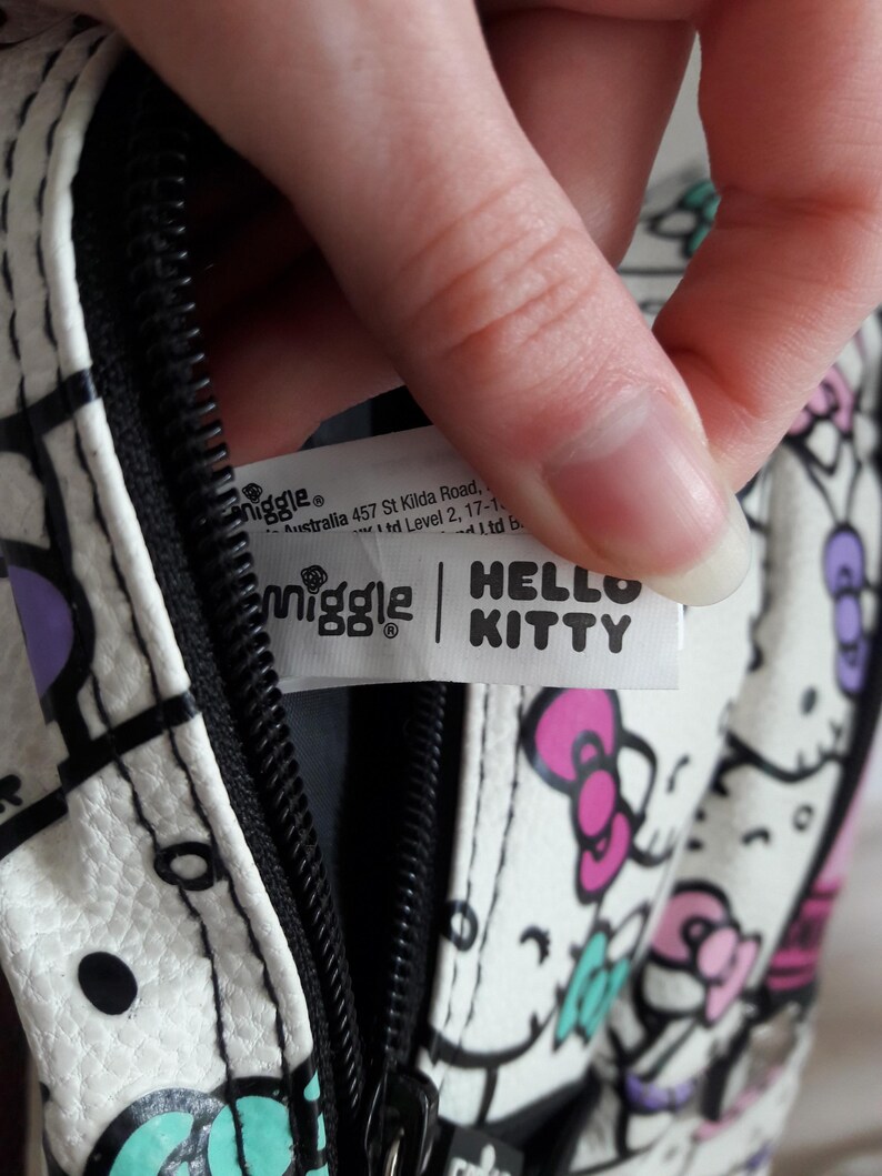 Puede incluir: Bolso blanco con cremallera negra y dise&ntilde;o de Hello Kitty. El bolso presenta un patr&oacute;n repetido de caras y lazos de Hello Kitty en rosa, morado y turquesa. El bolso tiene una etiqueta que dice "Smiggle" y "Hello Kitty."