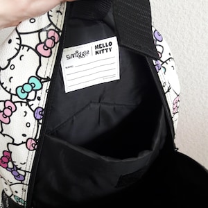 Puede incluir: Mochila blanca con un estampado repetido de caras de Hello Kitty con lazos rosas, morados y turquesas. La mochila tiene un interior negro y una etiqueta que dice "Smiggle | Hello Kitty". Tiene un asa negra y un bolsillo lateral de malla.