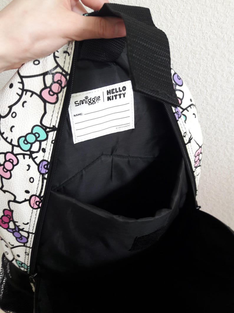 Puede incluir: Mochila blanca con un estampado repetido de caras de Hello Kitty con lazos rosas, morados y turquesas. La mochila tiene un interior negro y una etiqueta que dice "Smiggle | Hello Kitty". Tiene un asa negra y un bolsillo lateral de malla.