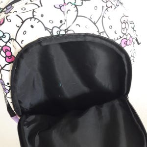 Puede incluir: Mochila blanca con un estampado repetido de caras de Hello Kitty con lazos rosas, morados y turquesas. La mochila tiene un interior negro y un bolsillo frontal con cremallera. La bolsa est&aacute; abierta, mostrando el interior negro.