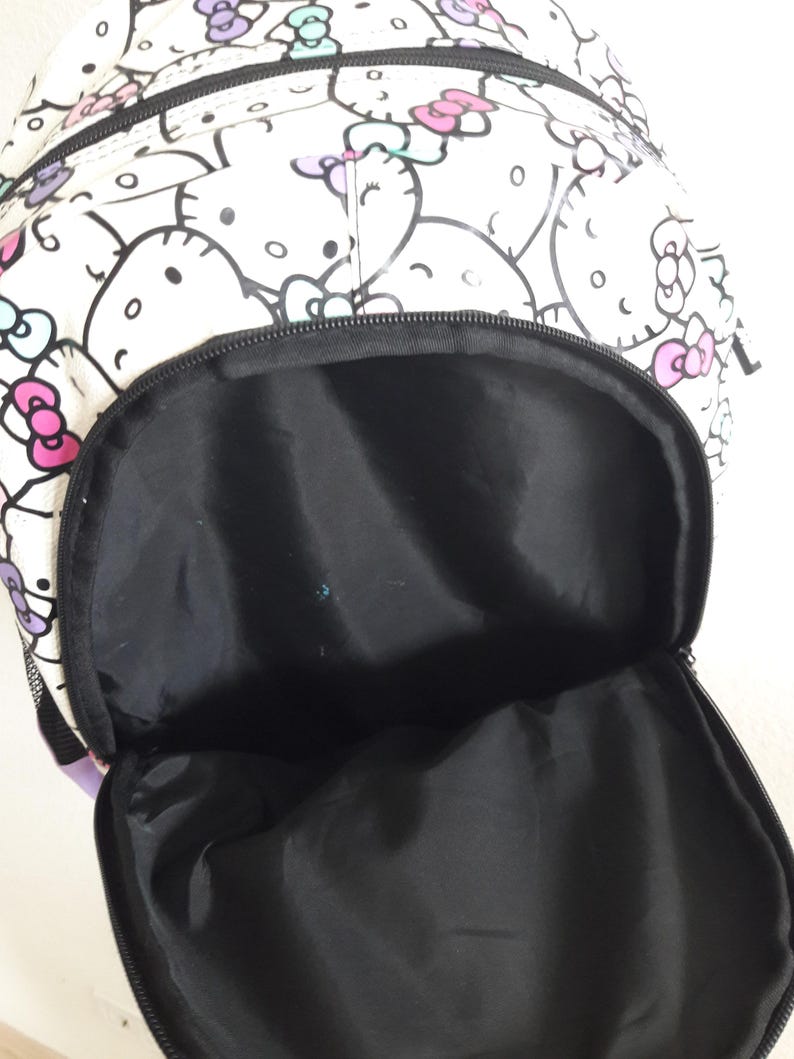 Puede incluir: Mochila blanca con un estampado repetido de caras de Hello Kitty con lazos rosas, morados y turquesas. La mochila tiene un interior negro y un bolsillo frontal con cremallera. La bolsa est&aacute; abierta, mostrando el interior negro.
